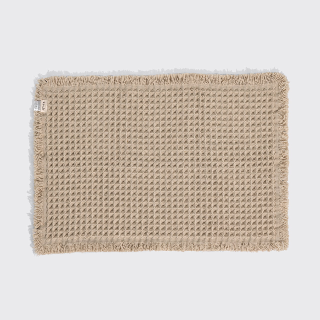 Camel waffle-knit cotton bath mat with fringed edges, laid flat, showing a Kitsch fabric tag on the left side.